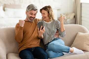 happy middle aged couple using smartphone shaking fists at home