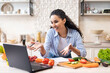 © Prostock-studio - Happy latin woman talking to laptop webcamera while preparing fresh salad in kitchen interior, having video call