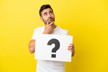 Young handsome caucasian man isolated on yellow background holding a placard with question mark symbol and thinking