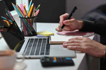 Poster - The close up of male using a pen to write something on a memo book on the desk with a computer laptop and stationery suppliers. Business and financial concepts.