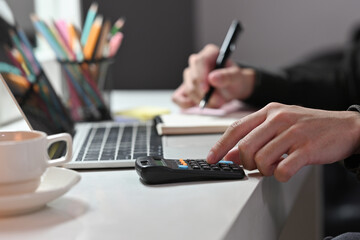 Poster - Close-up of male using a calculator and writing on a note next to a black screen laptop and a cup of coffee.Business and financial concepts.