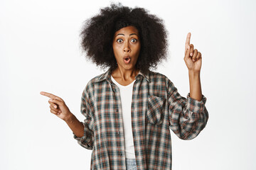 Wall Mural - Enthusiastic african female model with afro hair, pointing sideways, left and right, showing choices, store promo banners aside, standing over white background