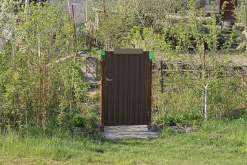 Poster - one closed brown metal door on a gray iron mesh fence wall on a rural street overgrown with green grass and vegetation