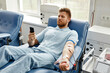 © Seventyfour - Portrait of young man donating blood while lying in comfortable chair at med center and using smartphone