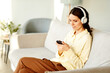 © Seventyfour - Young adult Caucasian woman wearing headphones sitting on couch at home choosing music to listen to on smartphone