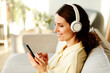 © Seventyfour - Portrait of attractive young adult Caucasian woman wearing wireless headphones sitting on sofa in living room listening to music and surfing Internet