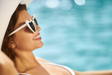 Beautiful Female In Sun Glasses Smiling, While Sunbathing Outdoors. Side View Of Happy, Young Woman Wearing White Hat, Lying On Sunbed, With Blurred Swimming Pool On Background. Concept Of Relaxation.