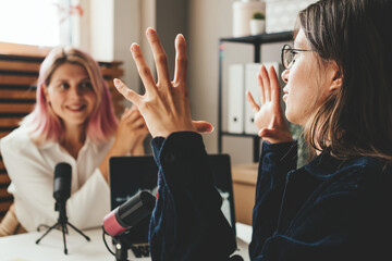 Two excited women host streaming audio podcast using microphone and laptop at home broadcast studio