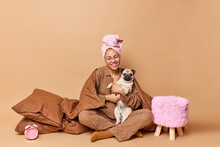 Domestic Atmosphere Friendship Between Animals And People. Cheerful Young European Woman Wears Bath Towel Wrapped On Head Embraces Pug Dog Sits Crossed Legs Under Blanket Enjoys Good Morning
