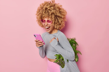 Wall Mural - Joyful woman with curly bushy hair uses mobile phone for making shopping online carries fabric bag full of healthy green vegetables for making vegetarian salad isolated over pink background.