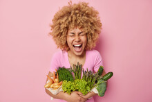 Joyful Curly Haired Woman Poses With Healthy Organic Food Full Of Vitamins Embraces Raw Fresh Vegetables Exclaims From Happiness Isolated Over Pink Background Going To Cook Vegetarian Salad.