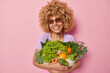 Cheerful woman wears sunglasses embraces bunch of fresh green vegetables picks up grocery from own garden looks gladfully away isolated over pink background going to prepares delicious meal.