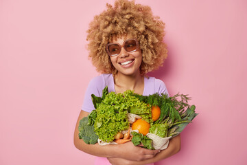 Wall Mural - Cheerful woman wears sunglasses embraces bunch of fresh green vegetables picks up grocery from own garden looks gladfully away isolated over pink background going to prepares delicious meal.