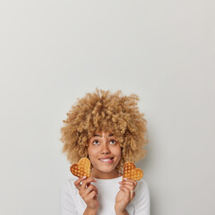 Vertical shot of curly haired woman bites lips focused above holds two heart shaped waffles eats appetizing dessert dressed in casual jumper isolated over white background blank space for text