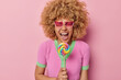 Overjoyed curly haired young woman laughs happily keeps mouth opened holds rainbow lollipop on stick exclaims from joy wears heart shaped sunglasses casual t shirt isolated over pink background