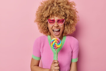 Wall Mural - Overjoyed curly haired young woman laughs happily keeps mouth opened holds rainbow lollipop on stick exclaims from joy wears heart shaped sunglasses casual t shirt isolated over pink background