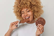 Portrait Of Cheerful Woman With Curly Hair Bites Appetizing Sweet Chocolate Holds Cookie Eats Favorite Desserts Isolated Over White Background. Unhealthy Harmful Food And Breaking Diet Concept