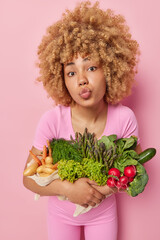 Wall Mural - Vertical shot of curly haired woman keeps lips rounded blows mwah poses with fresh green grocery eats vegetables keeps to diet isolated over pink background going to prepare vegeterian dinner