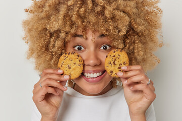Wall Mural - Close up portrait of cheerful woman with curly bushy hair holds cookies near face breaks diet has glad facial expression dressed in casual t shirt isolated over white background. Unhealthy food