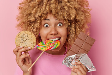 Wall Mural - Headshot of happy sweet tooth woman with curly bushy hair poses with favorite desserts holds cookie and bar of chocolate multicolored caramel lollipop in mouth isolated over pink background.