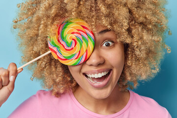 Wall Mural - Close up shot of cheerful curly haired young woman covers eye with big round multicolored caramel candy smiles broadly has fun dressed casually isolated over blue background. Sweet tooth concept