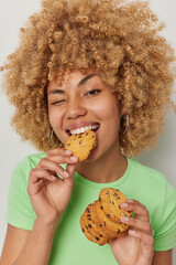 Wall Mural - Close up shot of positive curly haired young woman eats delicious appetizing cookies with chocolate winks eye dressed in casual green t shirt has sweet tooth addiction to sugar poses indoor.