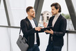 © anatoliycherkas - Two modern happy businessmen with documents folder discussing something against the background of urban offices and buildings