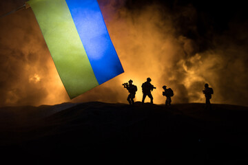 Russian war in Ukraine concept. Silhouette of armed soldiers against Ukrainian flag and burned out city. Creative artwork decoration. Night fighting scene