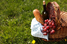 Wicker Basket With Picnic Blanket, Bottle Of Wine, Grapes And Bread On Green Grass. Space For Text