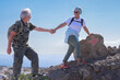 Active smiling senior couple in outdoor excursion hiking in mountain with backpack  enjoying healthy lifestyle. Woman helping man climb up. Horizon over water on background