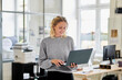 © Westend61 - Smiling young woman standing in office using laptop