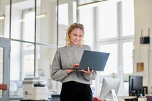 Smiling Young Woman Standing In Office Using Laptop