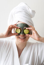 Smiling Young Woman With Face Mask Covering Eyes With Cucumber Slices In Front Of Wall