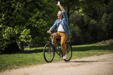 Smiling Senior Man Riding Bicycle Waving Hand At Park On Sunny Day
