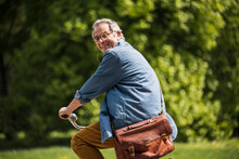 Happy Senior Man With Crossbody Bag Riding Bicycle At Park