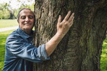 Smiling Senior Man Hugging Tree Trunk At Park