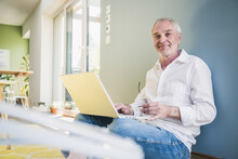 Smiling Senior Man With Laptop Leaning On Wall At Home