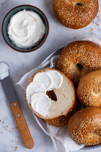 Bagel Cut In Half And Spread With Cream Cheese, Placed Inside Basket Full Of Sesame Bagels On White Marble Background