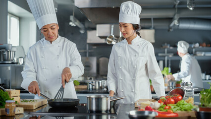 Wall Mural - Restaurant Kitchen: Portrait of Asian and Black Female Chefs Preparing Dish, Cutting Vegetables, Stirring Ingredients on the Frying Pan. Two Professionals Cook Delicious, Authentic Food, Healthy Meals