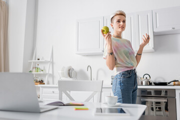 Wall Mural - smiling woman standing with apple and pen near blurred laptop and digital tablet on kitchen table