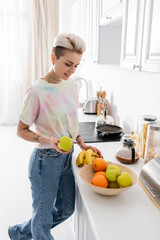 Wall Mural - trendy young woman holding apple and bananas near bowl with fresh fruits in kitchen