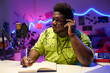 © AnnaStills - Portrait of stylish young African American musician sitting at desk listening to music track and working on lyrics