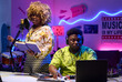 © AnnaStills - Young African American woman recording new song singing it into microphone, while her friend adjusting sound using laptop and mixer
