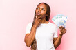 Young African American woman holding banknotes isolated on pink background looking sideways with doubtful and skeptical expression.