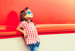 Summer portrait of little girl child wearing sunglasses, checkered shirt on red background in the city