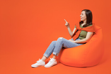 Full body young happy cheerful fun cool woman 20s wear khaki t-shirt sweater sit in bag chair point index finger aside on workspace area mock up isolated on plain orange background studio portrait.