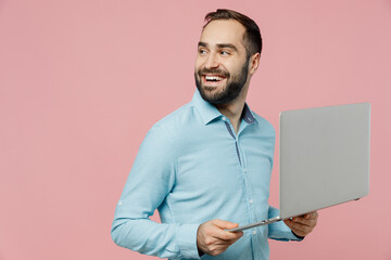 Young freelancer programmer caucasian man 20s wear classic blue shirt hold use work on laptop pc computer look aside on workspace area isolated on plain pastel light pink background studio portrait