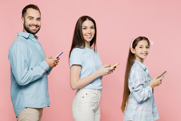 Wall Mural - Side view young happy parents mom dad with child kid daughter teen girl wear blue clothes hold in hand use mobile cell phone look camera isolated on plain pastel pink background. Family day concept