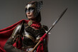 Shot of female barbarian holding spear dressed in steel armor with helmet against grey background.