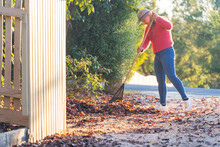 A Woman In A Bright Coloured Jumper Raking Up Leaves In A Driveway